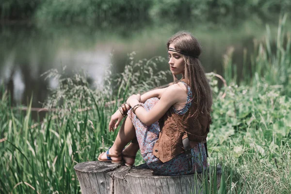Krásný hippie dívka sedí na pařezu poblíž rybníka. — Stock fotografie