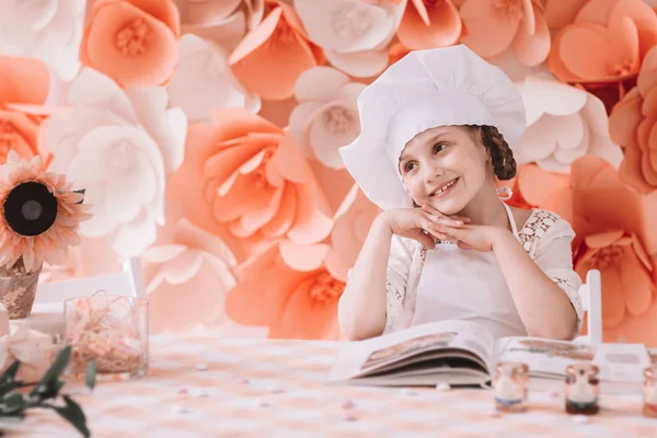 Очаровательная маленькая девочка в головной убор стоит за обеденным столом, готовая приготовить ужин. — стоковое фото