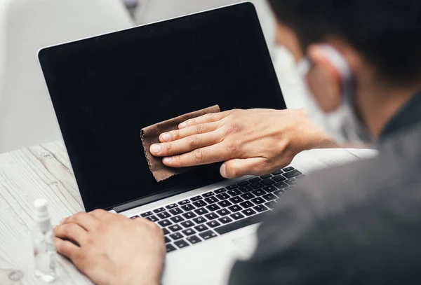 Nahaufnahme. ein junger Mann in Schutzmaske wischt den Bildschirm seines Laptops ab — Stockfoto