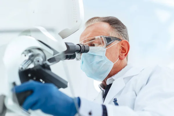 Científico en una máscara protectora mirando a través de un microscopio. — Foto de Stock