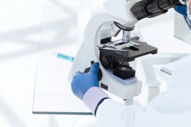 Koruyucu maskeli bir bilim adamı mikroskoptan bakıyor..