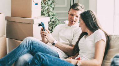 Akıllı telefonları olan genç bir çift yeni bir dairede kanepede oturuyor. .