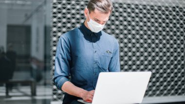 Koruyucu maskeli bir adam dizüstü bilgisayarın yüzeyine bakteriyel sprey sıkar..