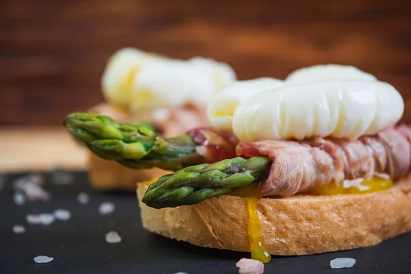 Grilled toast with asparagus in bacon and poached egg on wooden background