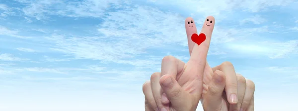 human hands with two fingers painted with red heart and smiley faces over cloudy blue sky background