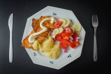 Meal Fried schnitzel with mashed potatoes, mayonnaise and tomato, onion and cucumber salad. On a dark background.