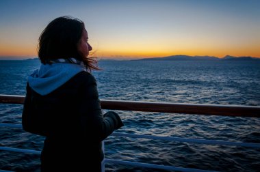 Bir yolcu gemisinin güvertesinden gün batımını izleyen kız.
