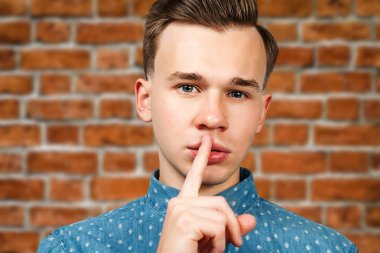Beyaz genç adam ağız yoluyla sessizlik hareketi onun işaret parmağı ile gösterilen mavi gömlek giymiş. Bir tuğla duvar arka plan üzerinde bir adam portresi