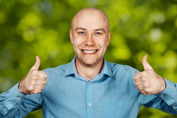 Portrait White bald guy in blue shirt on grass green background showing thumbs up and smiling
