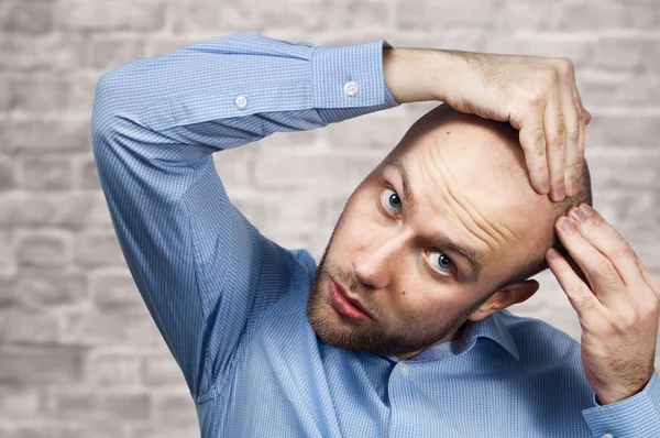 Portrait of a bald man looking at his balding head with a short haircut in a blue shirt. Hair transplant concept: white sad caucasian guy on Brick wall background