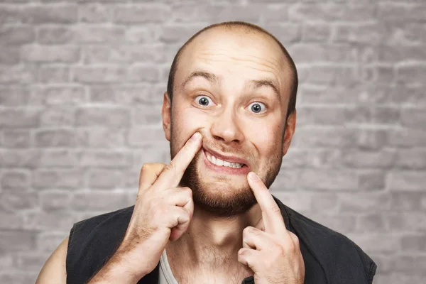 Retrato de cerca del joven calvo con barba, muecas, vestido con camisa sin mangas. Hombre sobre fondo de pared de ladrillo . — Foto de Stock