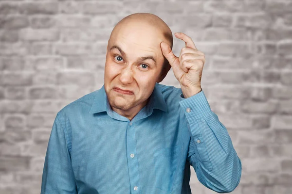 Portrait bald sad man thinks about money and a small salary. White bald guy in blue shirt on white brick wall background showing thumbs up small size.