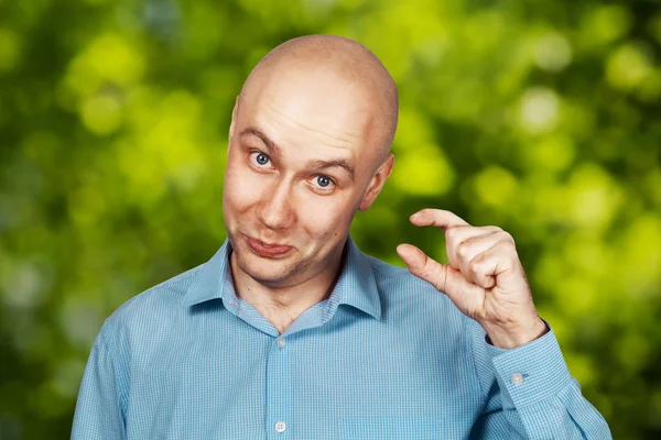 Portrait bald sad man thinks about money and a small salary. White bald guy in blue shirt on green bokeh background showing thumbs up small size.
