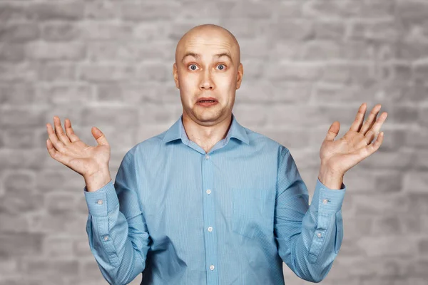 Retrato calvo hombre inseguro no entiende nada y extiende sus manos. chico en una camisa azul sobre fondo de pared de ladrillo blanco en la perplejidad — Foto de Stock