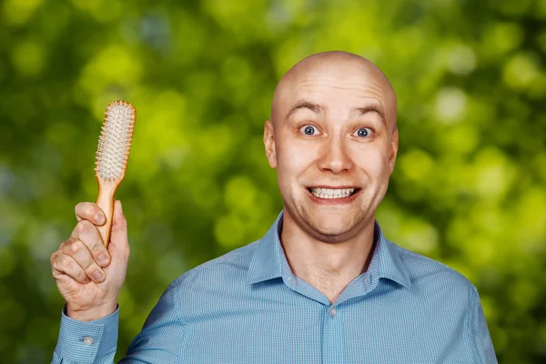 Portrait of a bald guy in a blue shirt holding a comb in his hands on green bokeh background. The concept of hair loss and hair transplantation