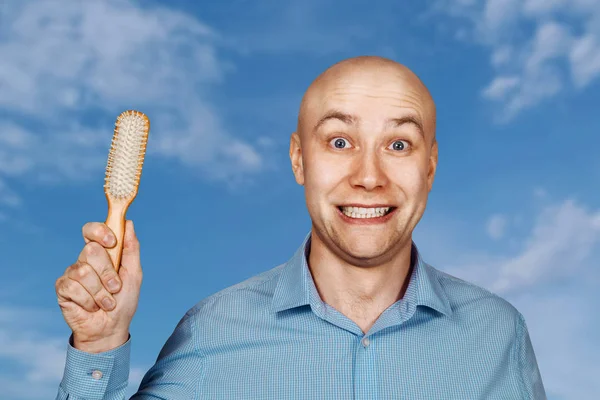 Portrait of a bald guy in a blue shirt holding a comb in his hands on blue sky background. The concept of hair loss and hair transplantation