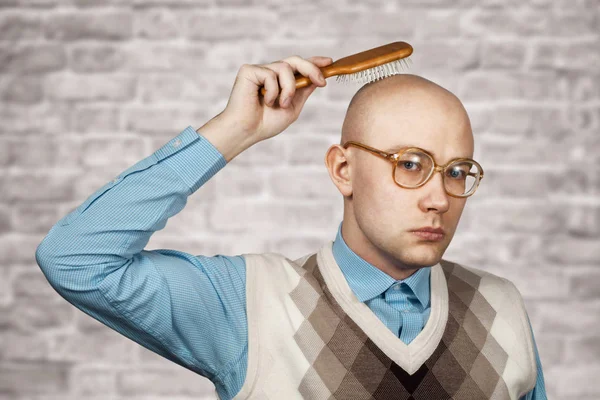 Portrait of a bald guy in a blue shirt holding a comb in his hands on brick wall white background. The concept of hair loss and hair transplantation