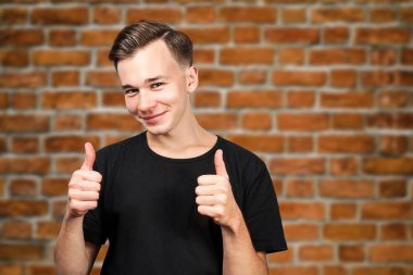 Siyah tişörtlü beyaz bir adam başparmak kaldırıyor, gülümsüyor ve duvara bakıyor.