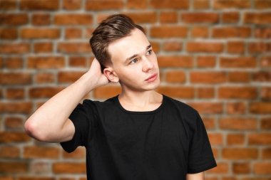 Siyah tişörtlü beyaz adam başının arkasında el ele tutuşup tuğla duvar arkasına bakıyor.