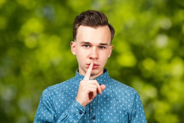Mavi gömlekli genç adam işaret parmağıyla sessizce hareket ediyor. Yeşil bokeh arka plandaki adam.