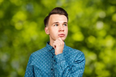 Mavi gömlekli beyaz adam başının arkasında el ele tutuşup yeşil bokeh arka planına bakıyor.