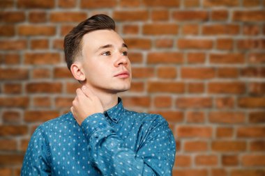 Mavi gömlekli beyaz adam başının arkasında el ele tutuşup tuğla duvar arkasına bakıyor.