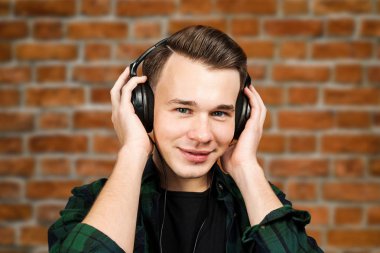 beyaz beyaz genç bir adamın portresi kulak telefonlarından müzik dinliyor. Yakışıklı adam siyah bir gömlek ve tuğla duvar arka plan üzerinde yeşil ekose gömlek giymiş