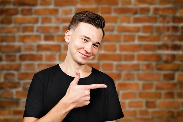 Vit ung kille klädd i svart t-shirt punkter åt sidan pekfinger och leenden på tegel vägg bakgrund — Stockfoto