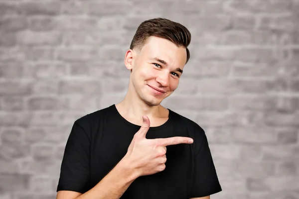 Fehér fiatal srác öltözött fekete póló pont félre mutatóujj és mosolyog téglafal háttér — Stock Fotó