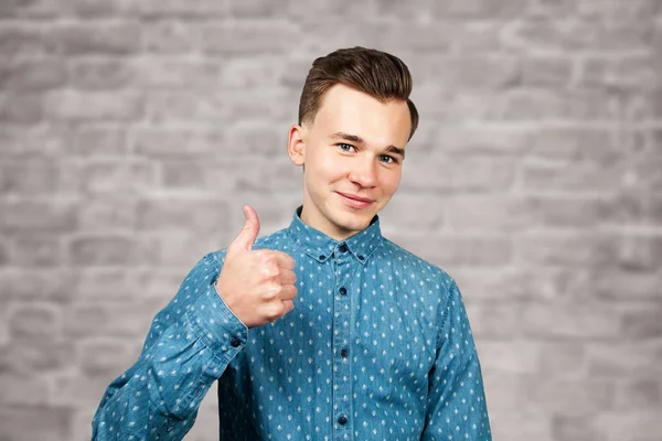 Λευκός νεαρός ντυμένος με μπλε πουκάμισο δείχνει τους αντίχειρες επάνω, χαμογελά και ψάχνει στο φόντο τούβλο τοίχο — Φωτογραφία Αρχείου