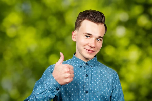 身穿蓝色衬衫的白人年轻人竖起大拇指，微笑着，看着绿色的Bokeh背景 — 图库照片