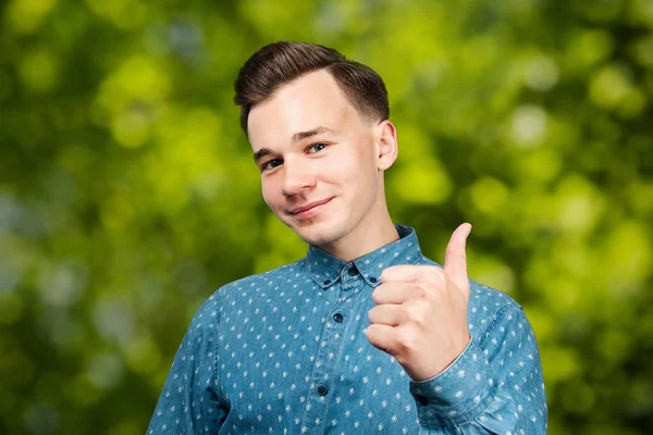 身穿蓝色衬衫的白人年轻人竖起大拇指，微笑着，看着绿色的Bokeh背景 — 图库照片
