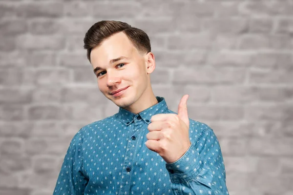 Λευκός νεαρός ντυμένος με μπλε πουκάμισο δείχνει τους αντίχειρες επάνω, χαμογελά και ψάχνει στο φόντο τούβλο τοίχο — Φωτογραφία Αρχείου