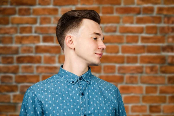 White young guy dressed in blue shirt on brick wall background smiles and looking aside — 스톡 사진