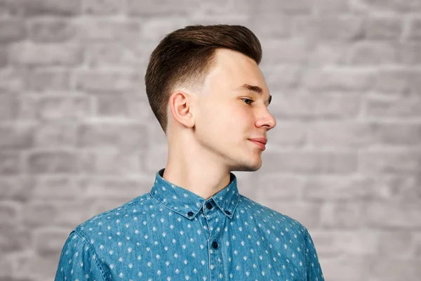 Fehér fiatal srác öltözött kék ing téglafal háttér mosolyog, és félrenéz — Stock Fotó