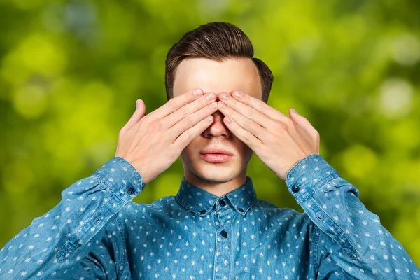 Πορτρέτο λευκός νέος άντρας ντυμένος με μπλε πουκάμισο, κλείνει τα μάτια στα χέρια του. Ο άνθρωπος στο πράσινο bokeh φόντο — Φωτογραφία Αρχείου