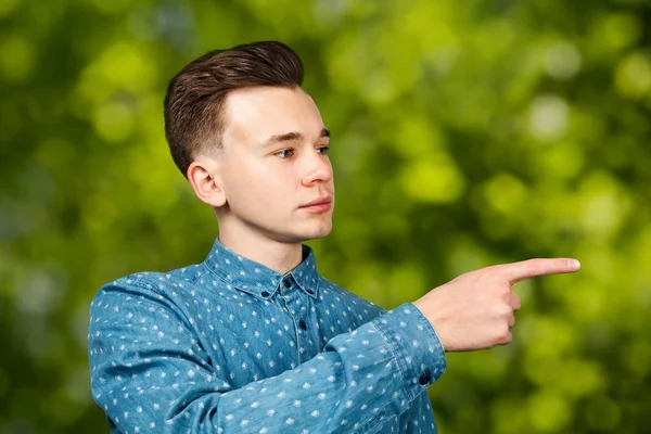 Joven blanco vestido con camisa azul apunta a un lado dedo índice sobre fondo bokeh verde — Foto de Stock