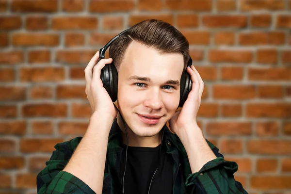 Porträtt av en kaukasisk vit ung kille lyssna musik i örat-telefoner. Stilig man klädd i en svart skjorta och en grön pläd skjorta på tegelvägg bakgrund — Stockfoto