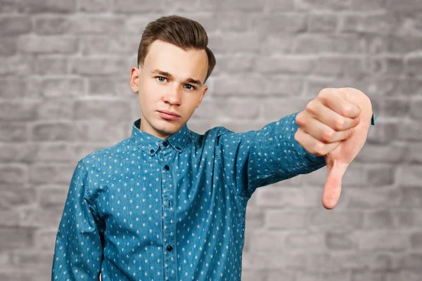 White young man dressed in blue shirt showing thumbs down on brick wall background