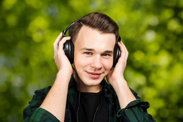 Πορτρέτο ενός καυκάσιου λευκού νεαρού που ακούει μουσική στα ακουστικά. Όμορφος άντρας ντυμένος με μαύρο μπλουζάκι και πράσινο καρό πουκάμισο στο πράσινο bokeh φόντο — Φωτογραφία Αρχείου