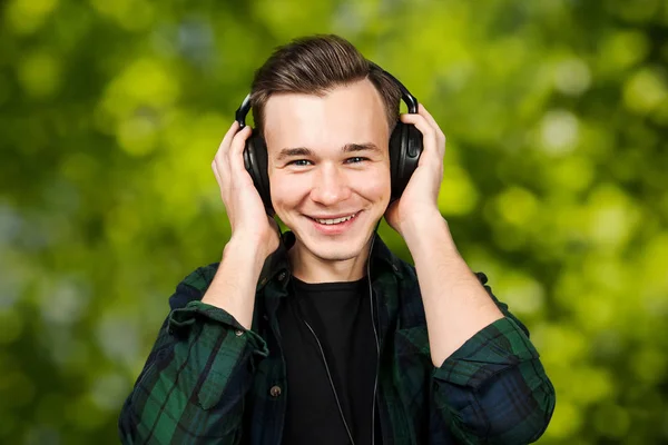 Retrato de um branco branco jovem ouvir música nos fones de ouvido. Bonito homem vestido com uma camiseta preta e uma camisa xadrez verde no fundo bokeh verde — Fotografia de Stock