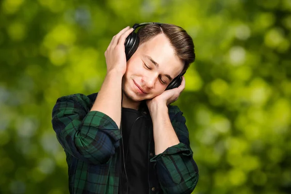 Porträtt av en kaukasisk vit ung kille lyssna musik i örat-telefoner med slutna ögon. Snygg man klädd i en svart T-shirt och en grön rutig skjorta på grön bokeh bakgrund — Stockfoto