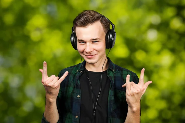 Retrato de um branco branco jovem ouvir música no fone de ouvido rock. Bonito homem vestido com uma camiseta preta e uma camisa xadrez verde no fundo bokeh verde — Fotografia de Stock