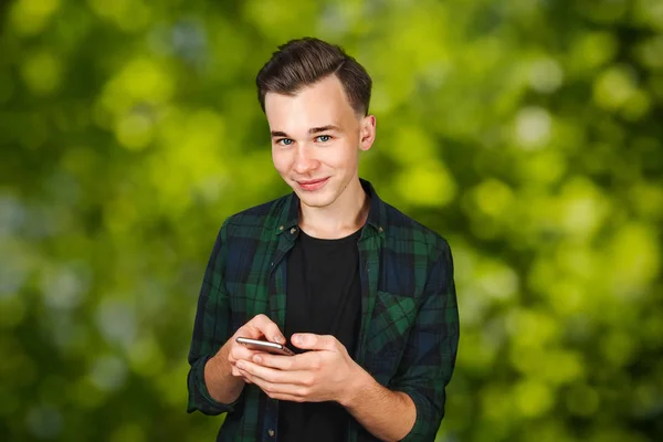 Egy fehér fiatalember üzenetet ír a mobiljára, és mosolyog. Férfi zöld bokeh háttér — Stock Fotó