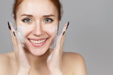 Neşeli gülen kız yüzünde yıkama için köpük uygulayan portresi. Çekici bir görünüme sahip güzel kadın kızıl saçlı. Gri arka planda Cilt Bakımı spa dinlenmek kavramı