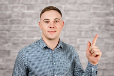 Mavi gömlek giymiş yakışıklı genç adam Portresi işaret parmağı yukarı gösterir ve tuğla duvar arka plan üzerinde gülümsüyor