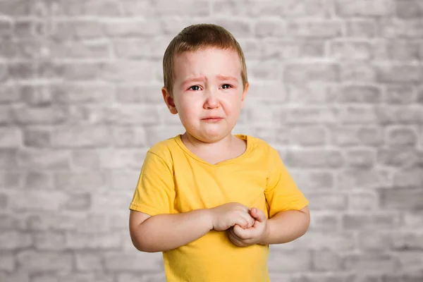 Crying baby boy in a yellow T shirt covers his face with hands and shouts, studio on brick wall background — Stock Photo, Image