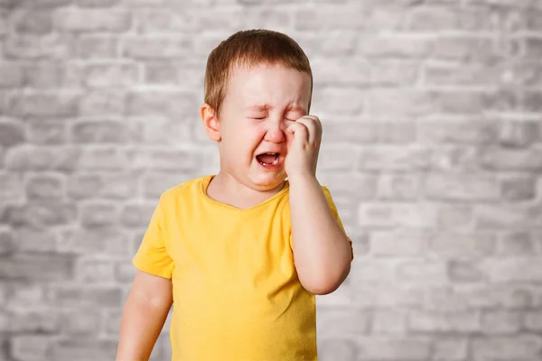 Chorando menino em uma camiseta amarela cobre seu rosto com mãos e gritos, estúdio no fundo da parede de tijolo — Fotografia de Stock