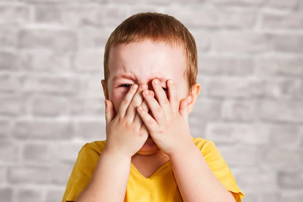Crying baby boy in a yellow T shirt covers his face with hands and shouts, studio on brick wall background — Stock Photo, Image