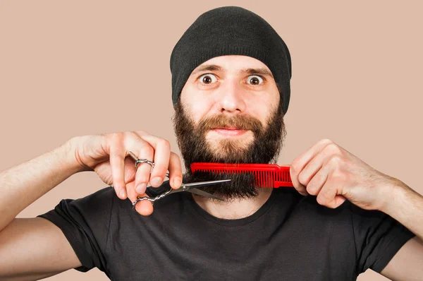 Bebaarde jonge kerel in hoed met kam en schaar snijden zijn baard. Geïsoleerd op bruine achtergrond — Stockfoto